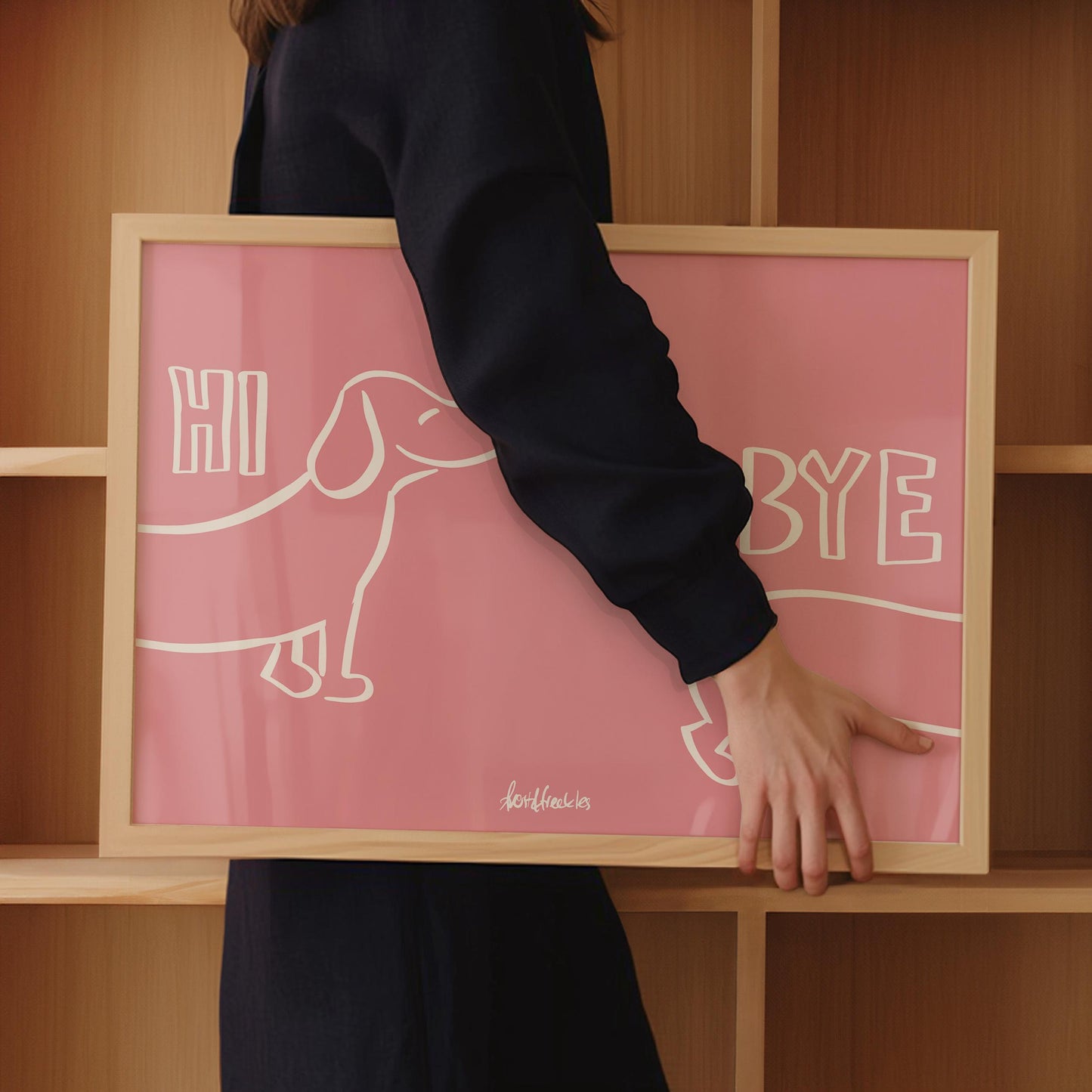 A woman in dark clothing holds a framed pink print with Dachshund illustration and the words "Hi Bye", in front of wooden shelves.
