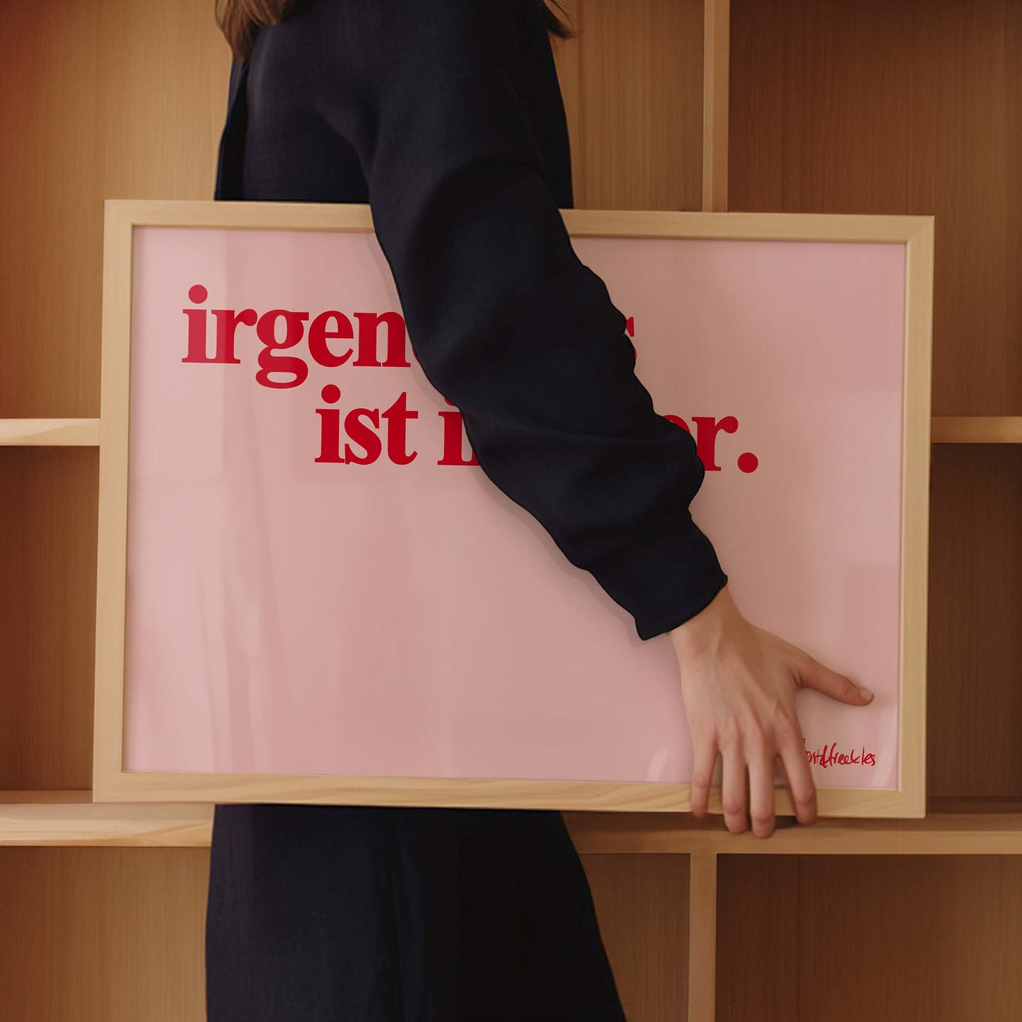 Eine Frau hält einen Bilderrahmen unter dem Arm, das Poster zeigt den Spruch "irgendwas ist immer" in roter Schrift auf pinkem Hintergrund.