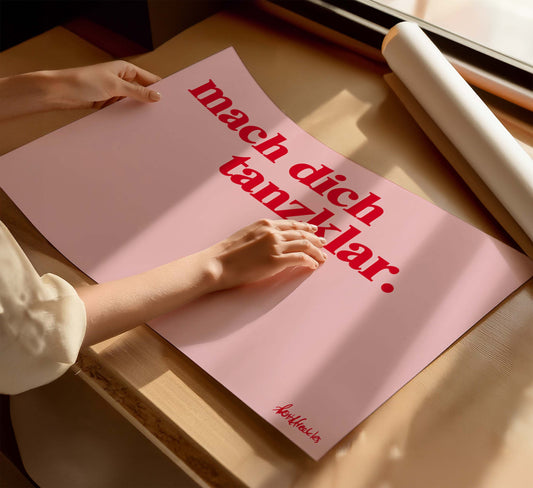 Eine Frau hält ein Poster mit dem deutschen Spruch "Mach dich Tanzklar", das Poster liegt flach auf einem Tisch neben einer Posterrolle.