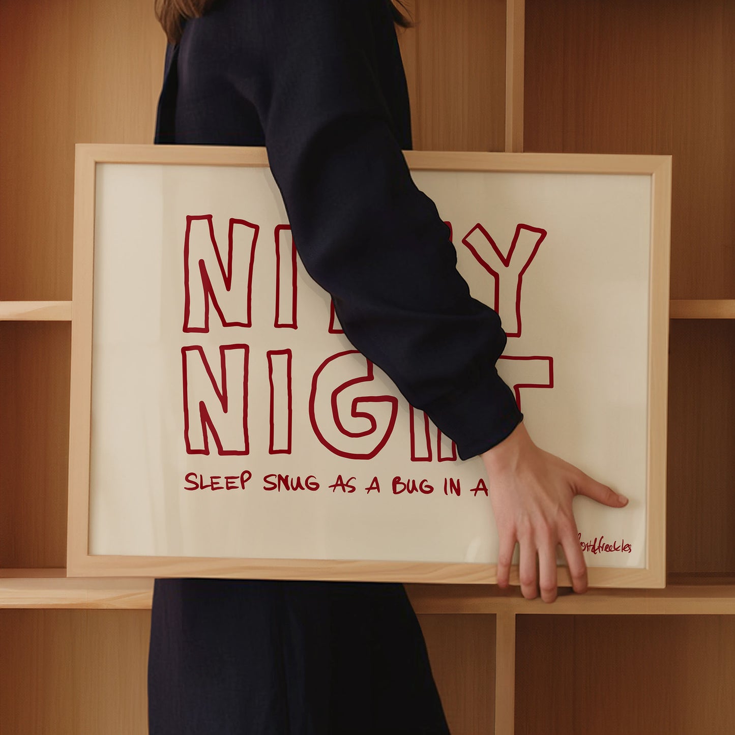 A woman in dark clothing holds a framed beige print with red text: "Nitey Night", and a smaller text "Sleep Snug As A Bug In A Rug", in front of wooden shelves.