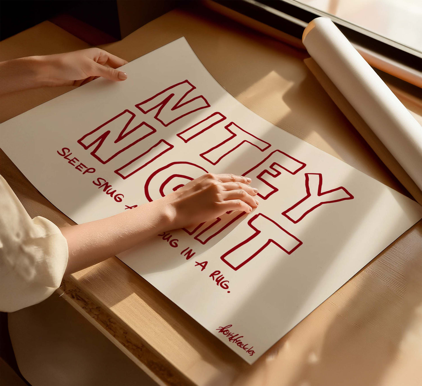 A woman holding a beige red poster with the phrase “Nitey Night", and subtitle "Sleep Snug As A Bug In A Rug", on a wooden table near a window with sunlight.
