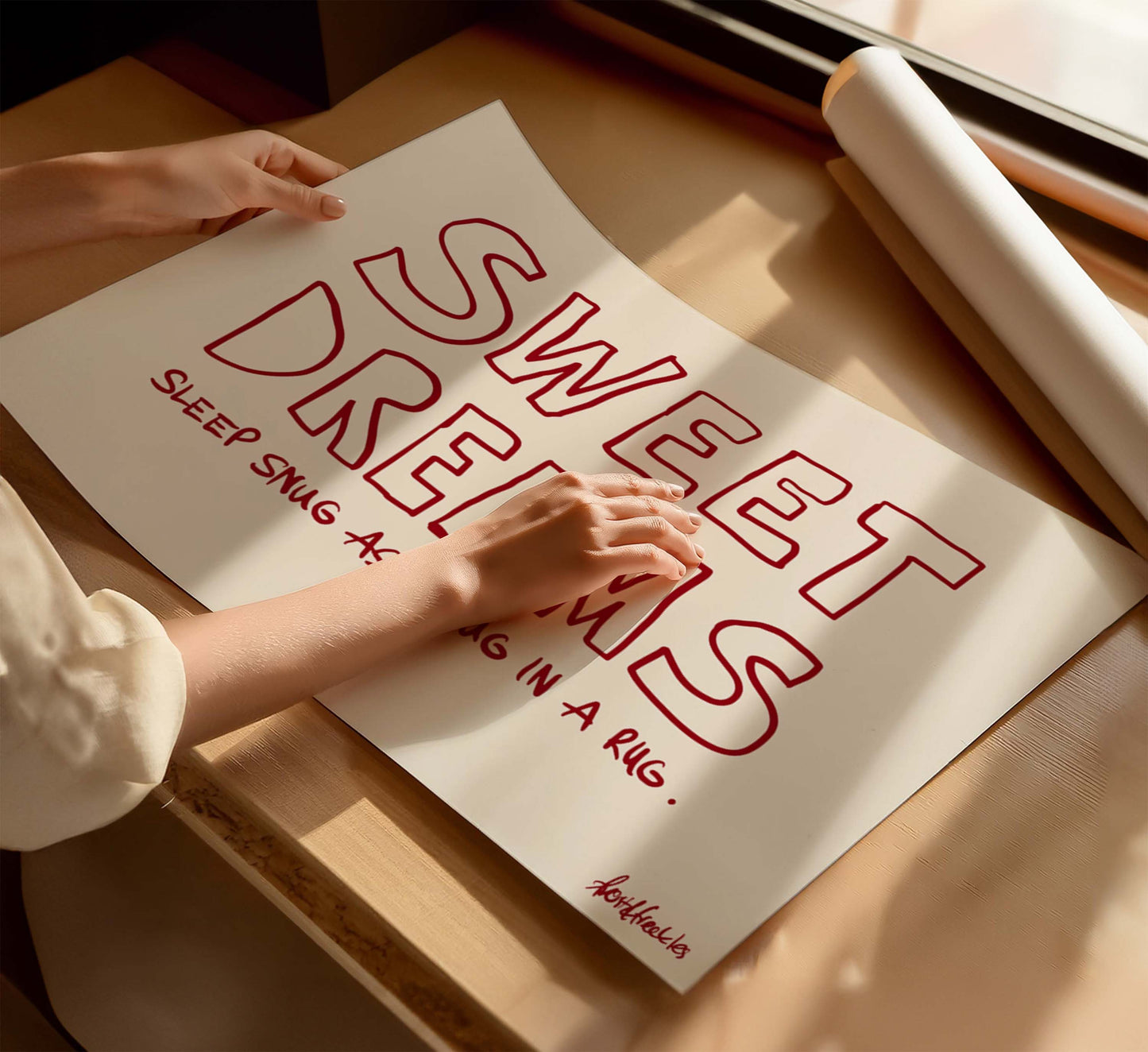 A woman holding a beige poster with the phrase “Sweet Dreams", in bold red text, on a wooden table near a window with sunlight.