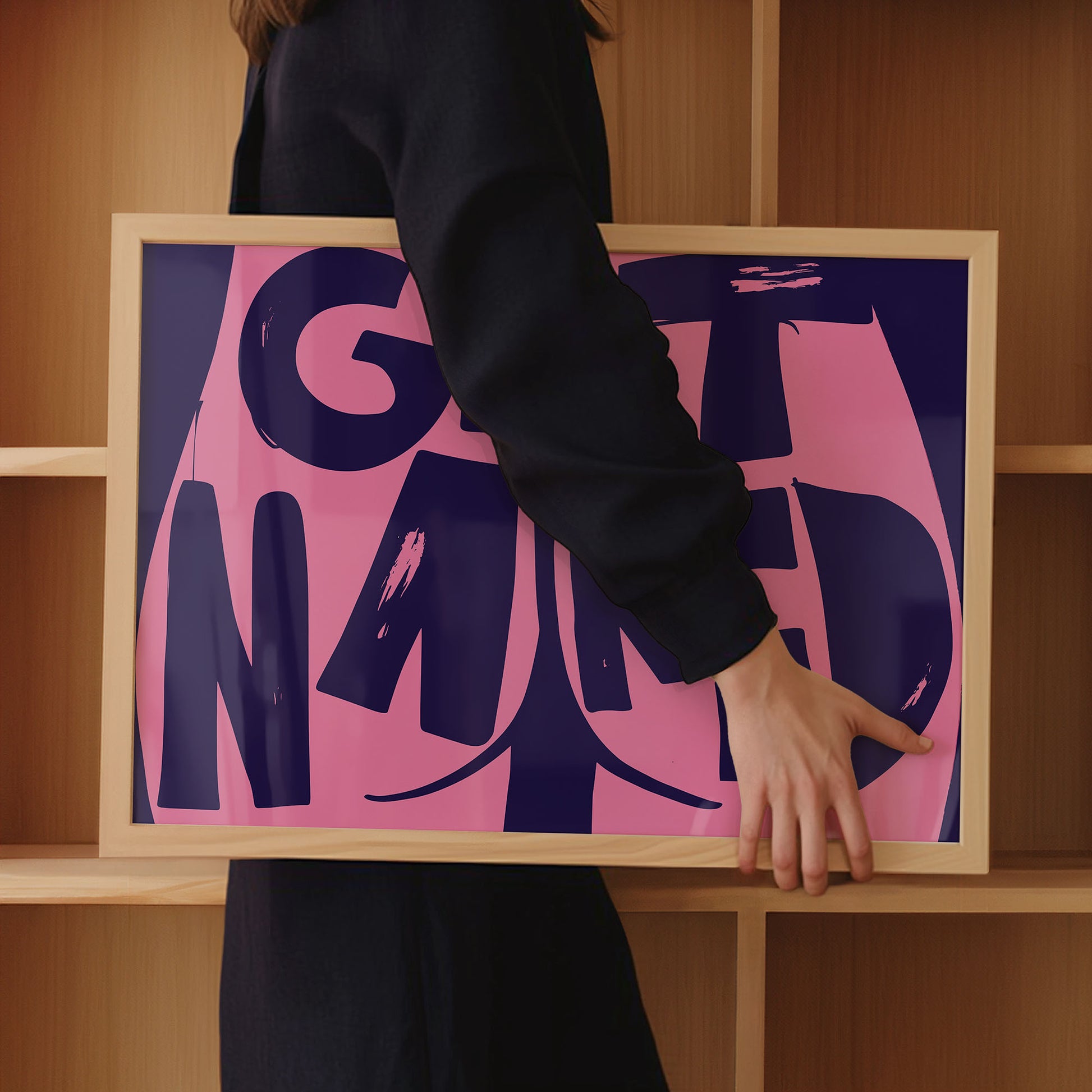 A woman in dark clothing holds a framed pink and purple print with the text: "Get Naked", alongside a butt cheek illustration, in front of wooden bathroom shelves.