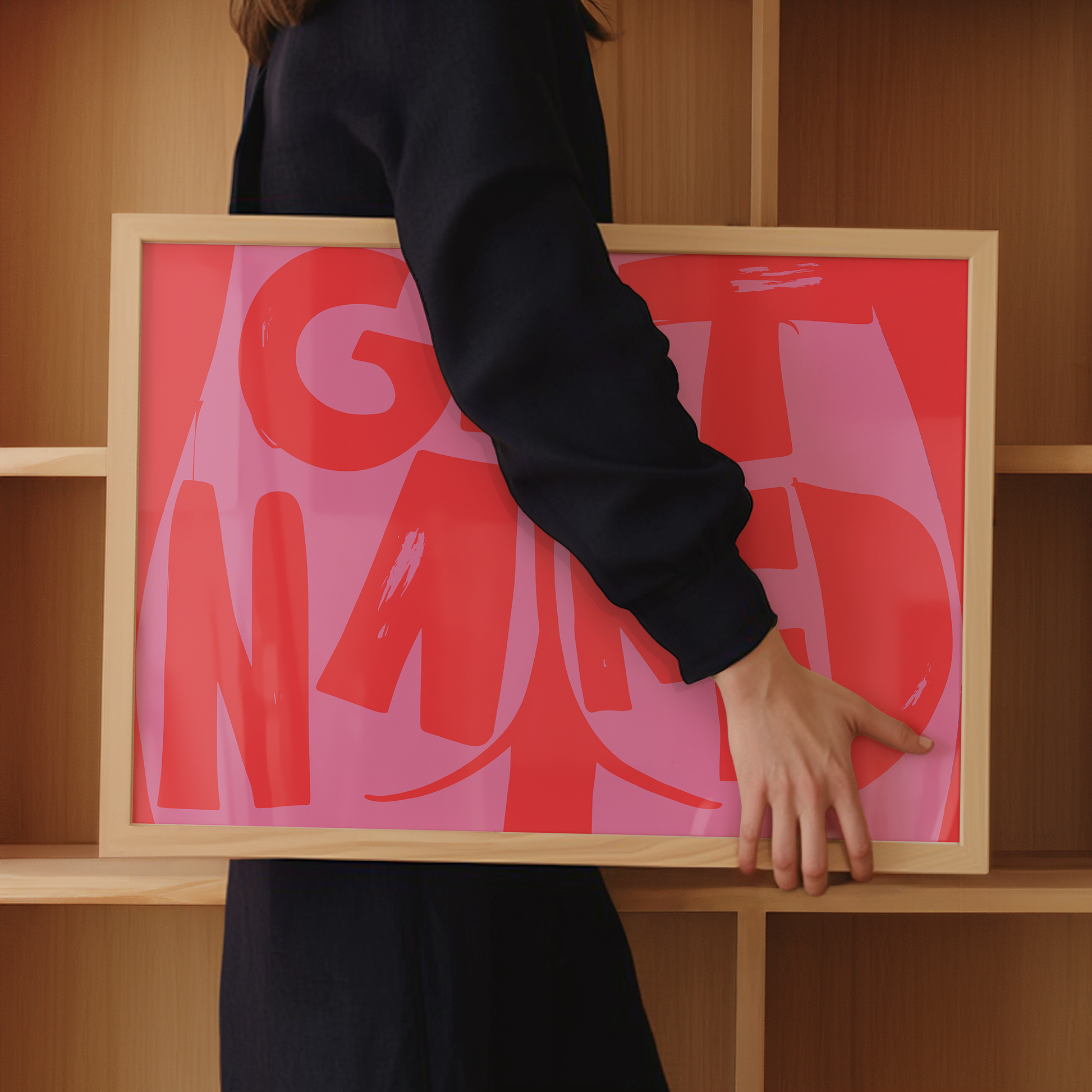 A woman in dark clothing holds a framed pink print with the text: "Get Naked", alongside a butt cheek illustration, in front of wooden shelves.
