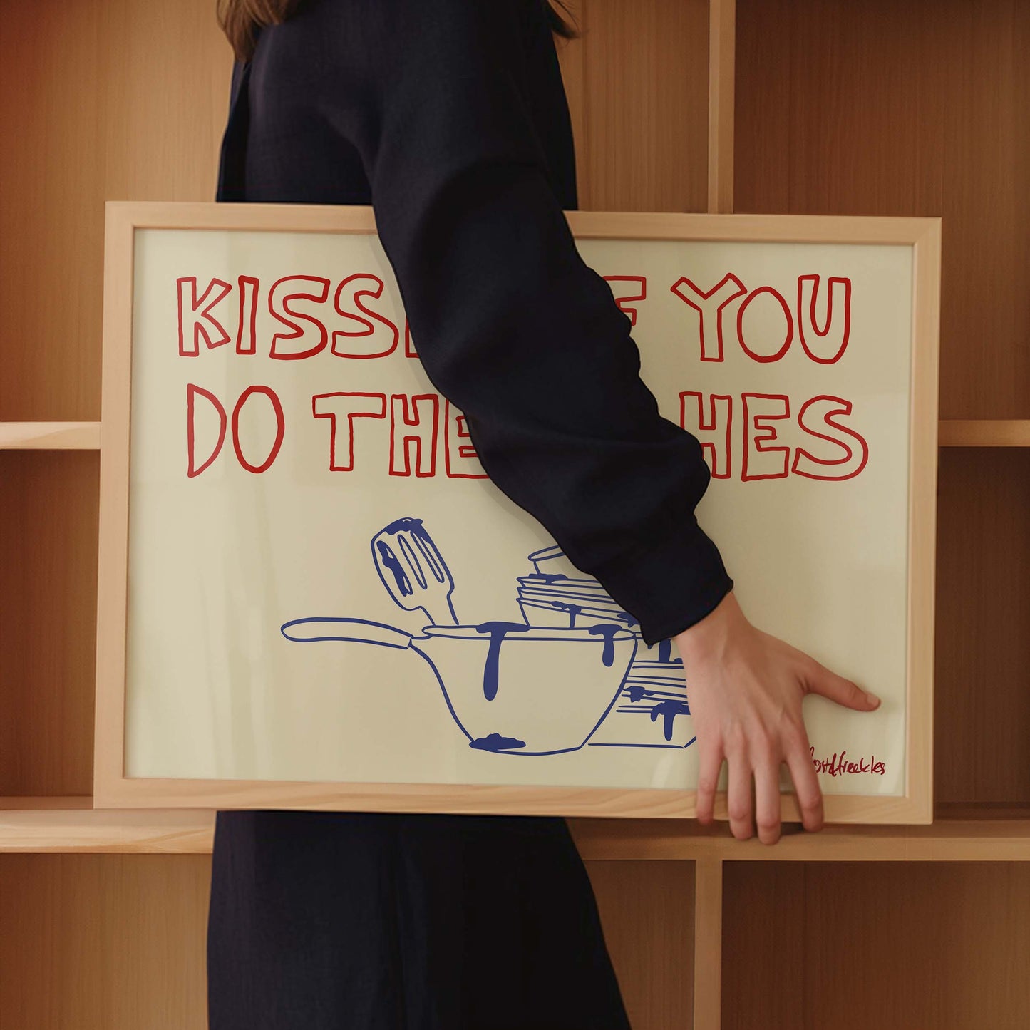 Cheeky wall print with handwritten font reading "Kisses If You Do The Dishes" on beige background next to open shelving