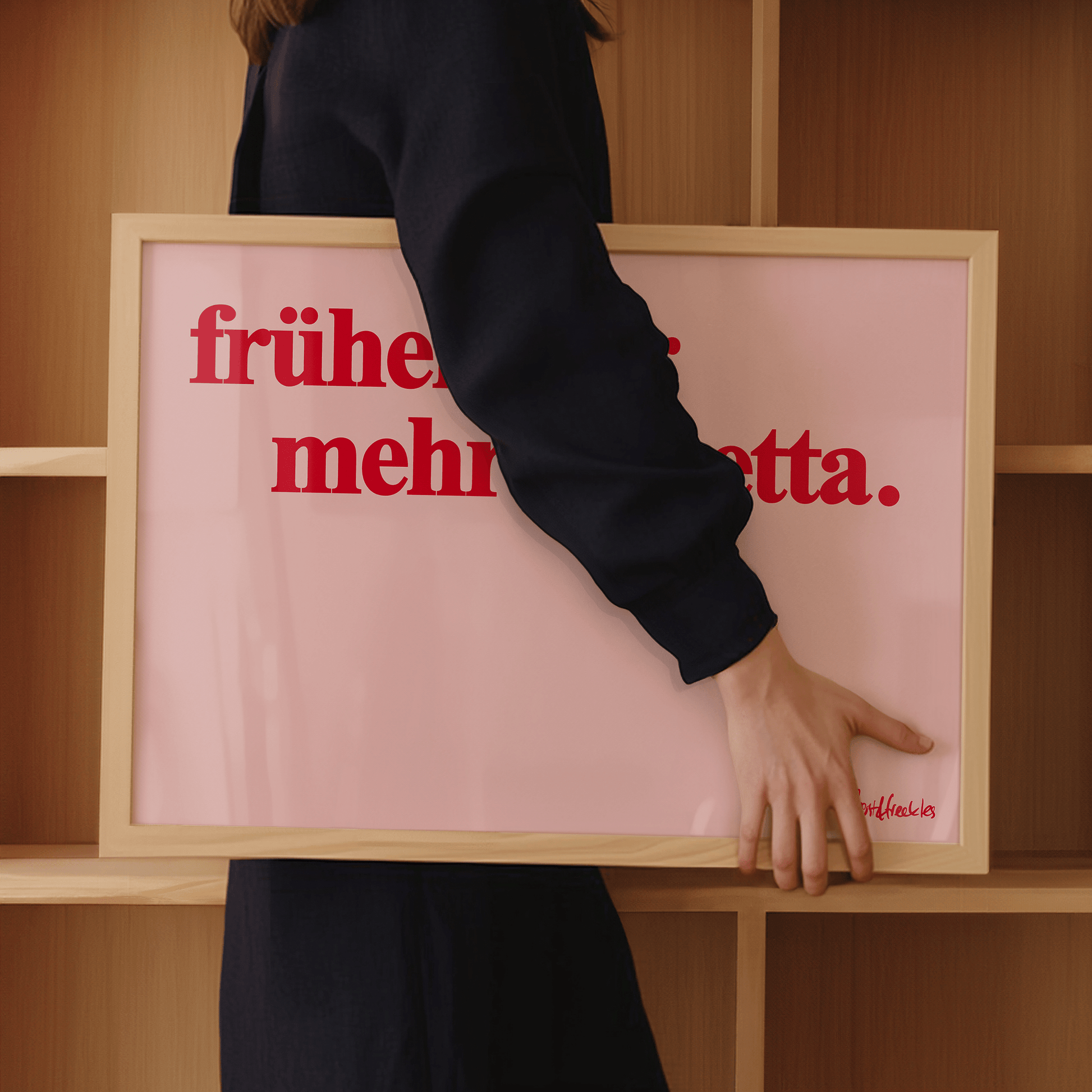 A woman in dark clothing holds a framed pink print with red German text: "Früher mehr Lametta", a quote by Loriot, in front of wooden shelves.
