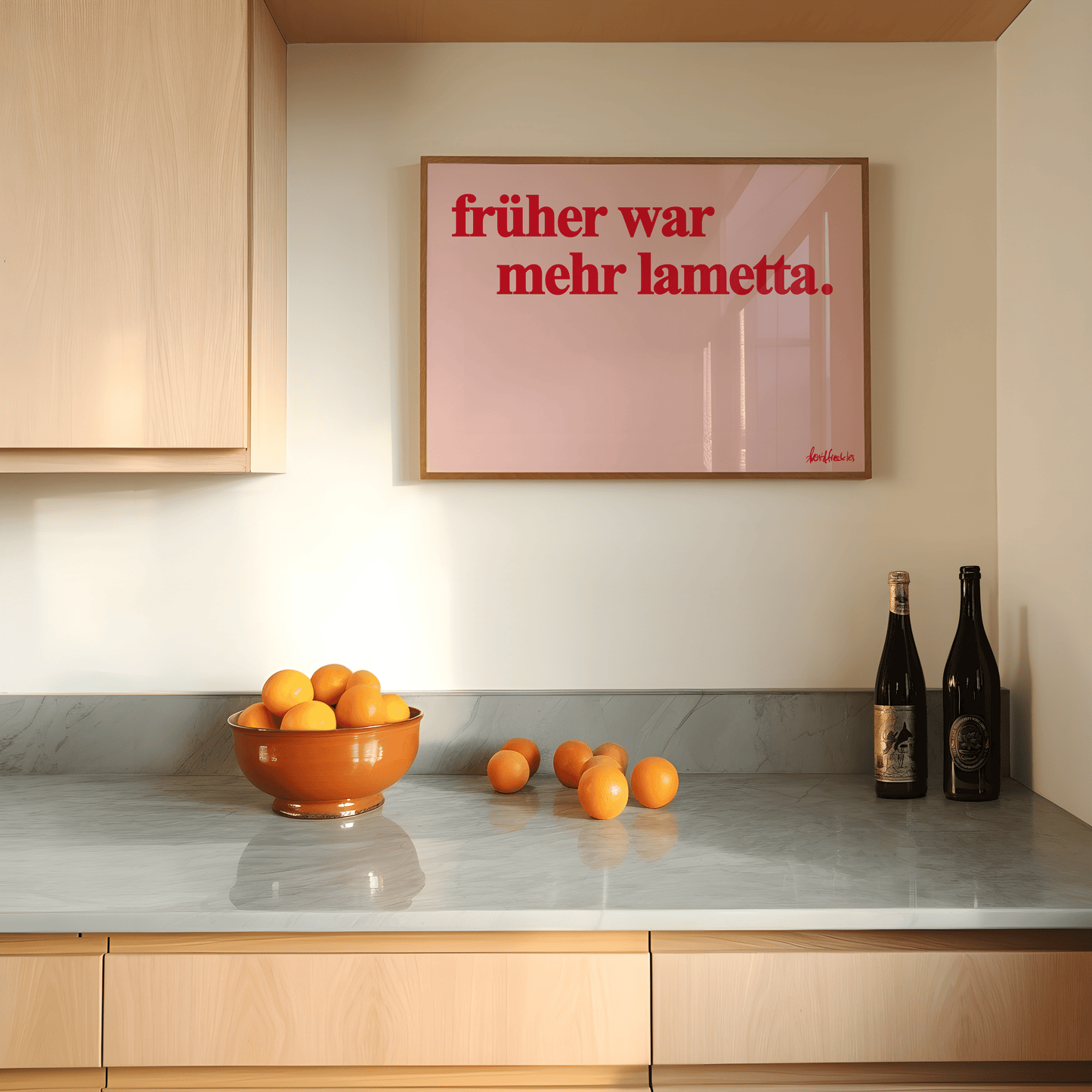 A modern kitchen counter with a bowl of oranges, loose oranges, two wine bottles, and a framed sign on the wall that reads "Früher war mehr Lametta" - a German quote by Loriot.