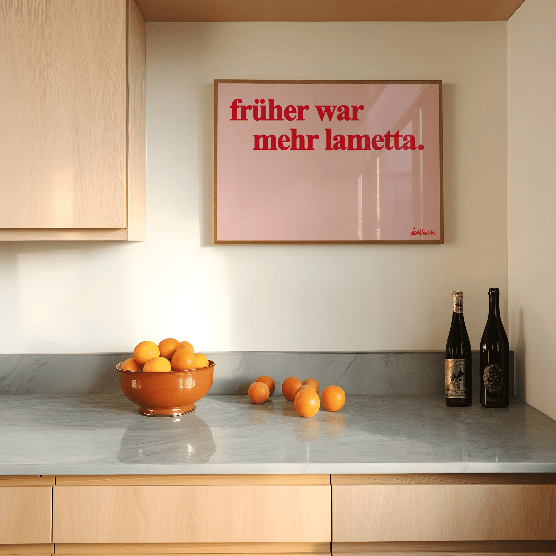 A modern kitchen counter with a bowl of oranges, loose oranges, two wine bottles, and a framed sign on the wall that reads "Früher war mehr Lametta" - a German quote by Loriot.