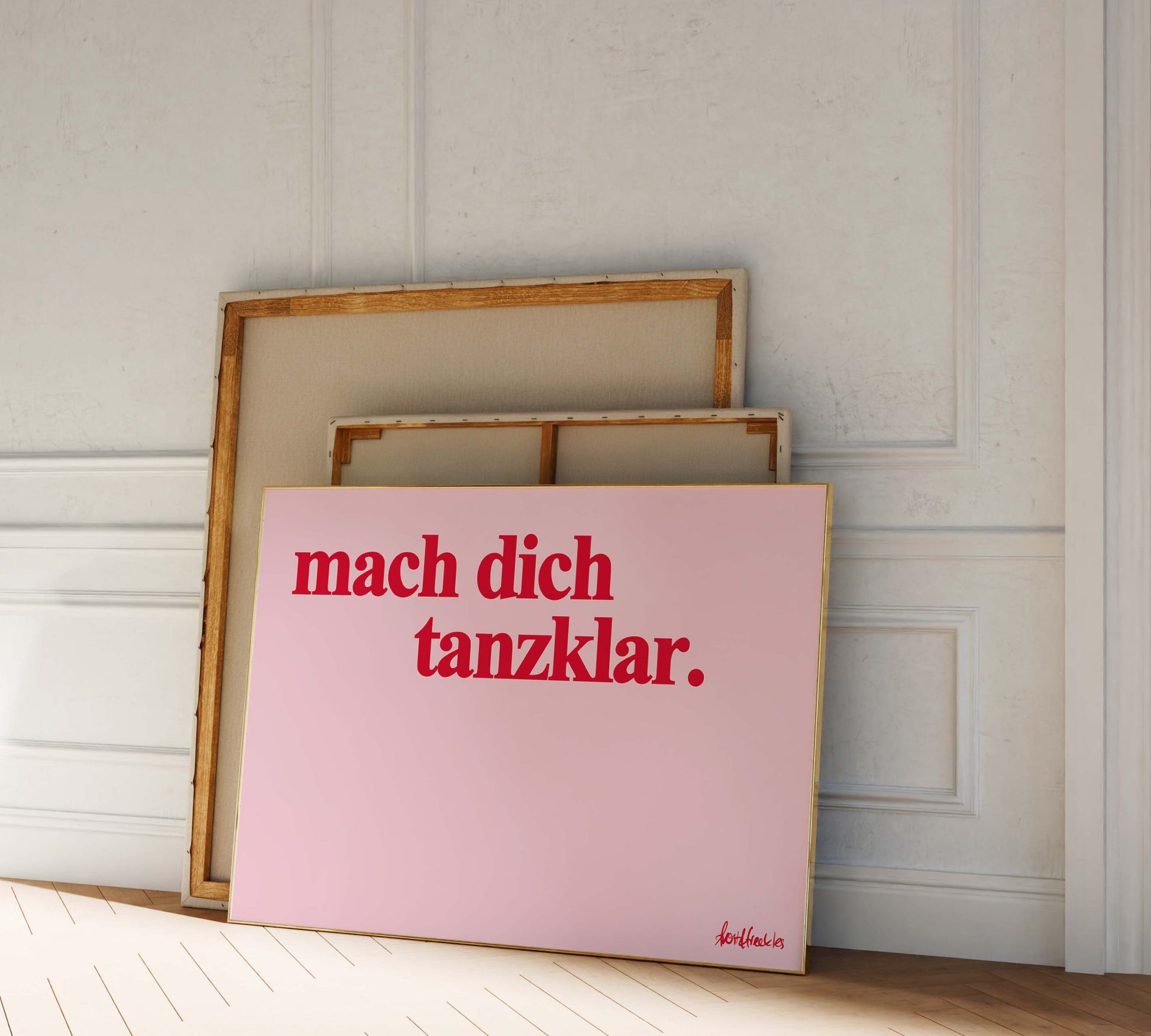 Zwei Bilderrahmen lehnen an einer Wand, das vordere Bild zeigt den deutschen Spruch "mach dich tanzklar" in roter Schrift auf rosé Hintergrund.