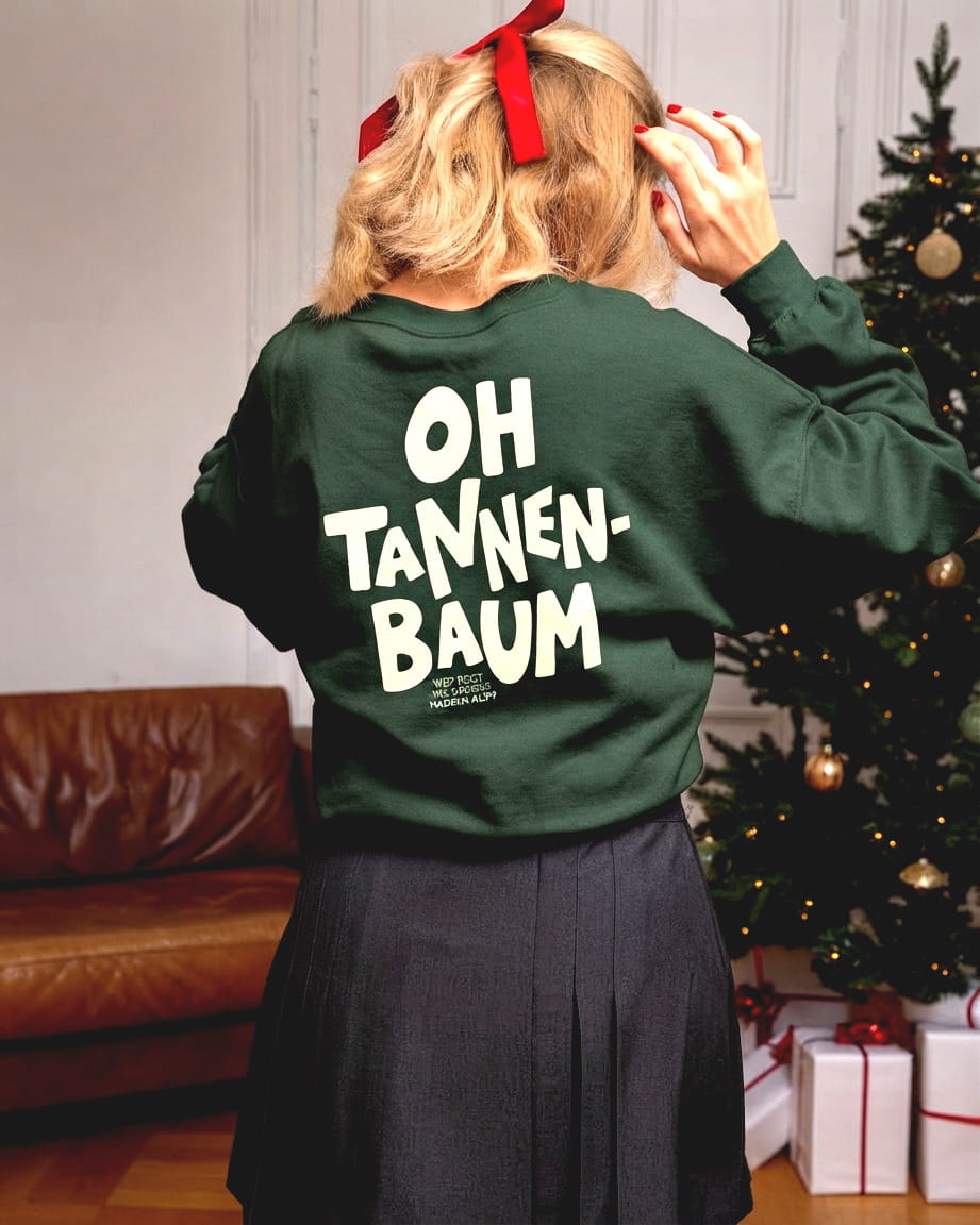 Frau trägt den lustigen "Oh Tannenbaum" Oversized Sweater in Forest Green unter dem Weihnachtsbaum mit einer festlichen Schleife im Haar.