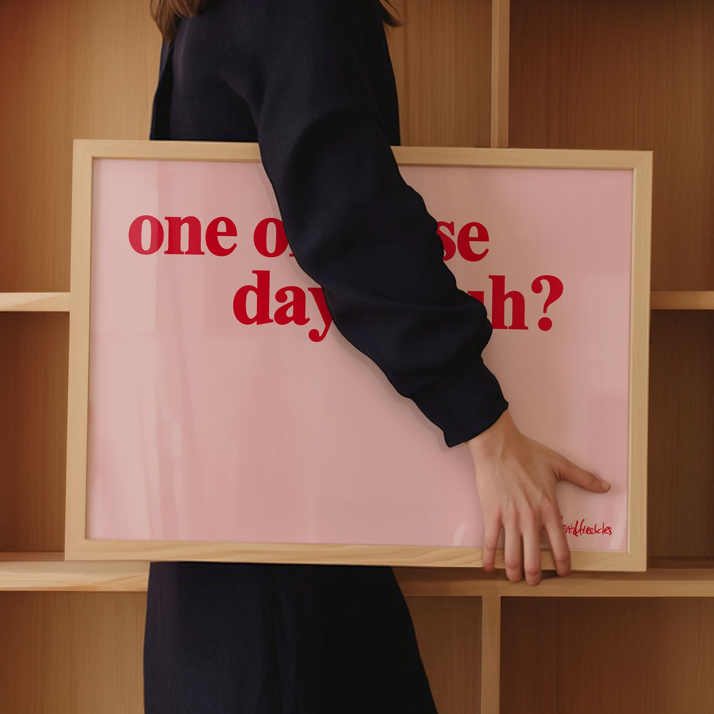 A woman in dark clothing holds a framed pink print with red text: "one of those days", in front of wooden shelves.

