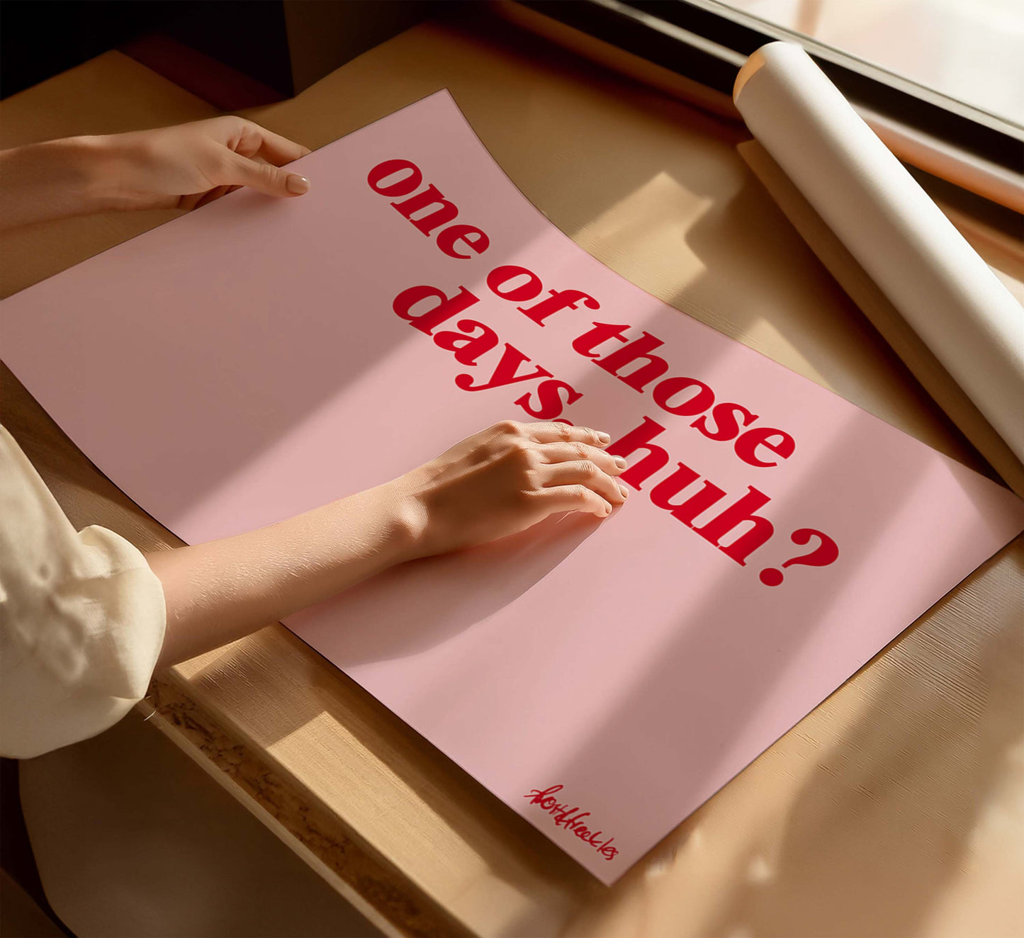 A woman holding a pink poster with the funny phrase “one of those days", in bold red text, on a wooden table near a window with sunlight.

