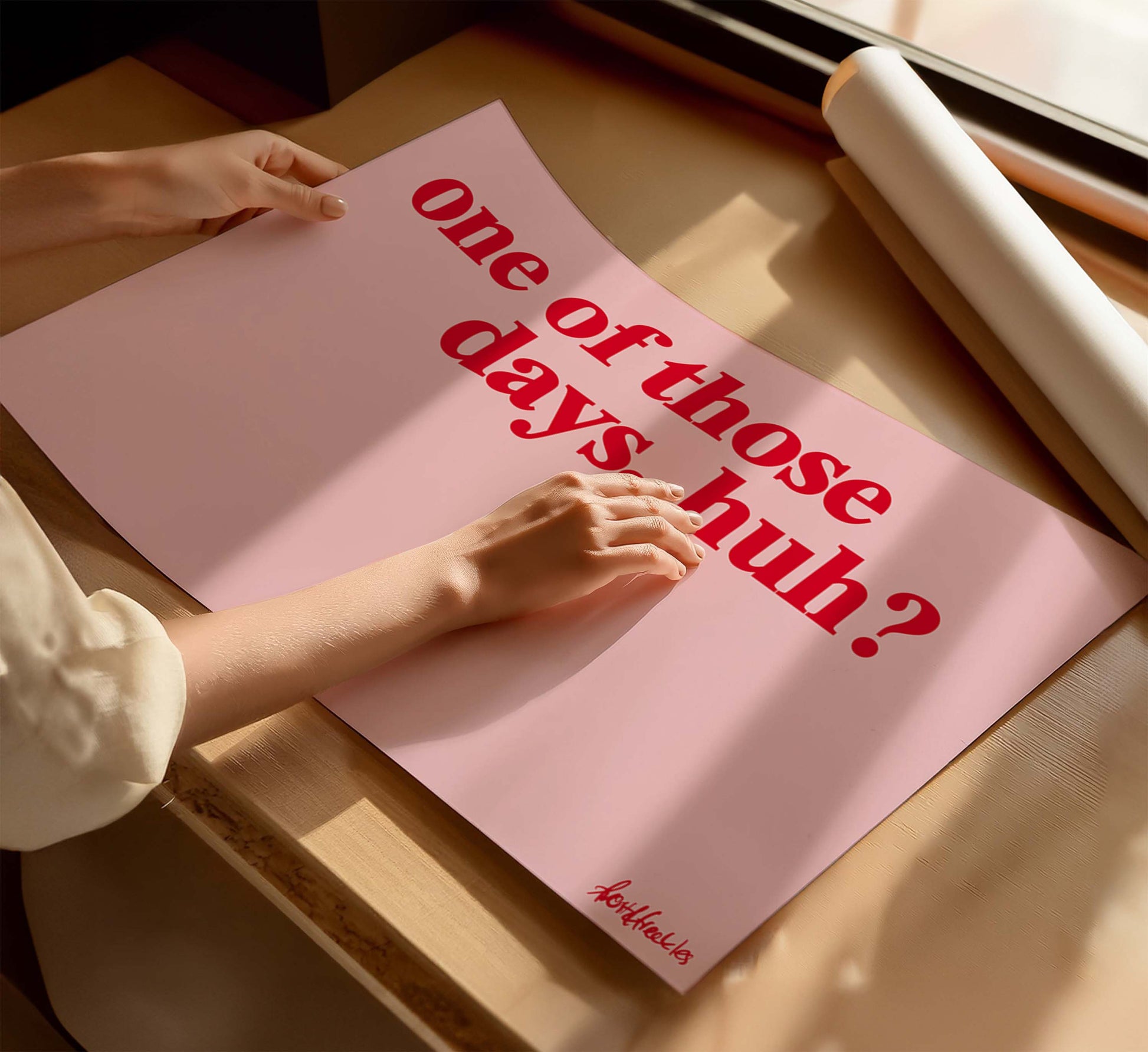 A woman holding a pink poster with the funny phrase “one of those days", in bold red text, on a wooden table near a window with sunlight.
