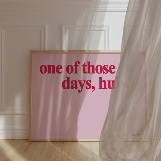 A framed picture leaning against a bedroom wall, pink background and bold red letters reading "one of those days huh", wind blowing the curtain around.