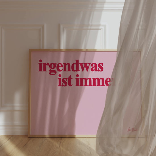 Ein gerahmtes, horizontales Poster zeigt den Spruch "Irgendwas ist immer" in roter Schrift auf pinkem Hintergrund. Der Wind weht einen Vorhang vor den Rahmen und verdeckt diesen teilweise. 