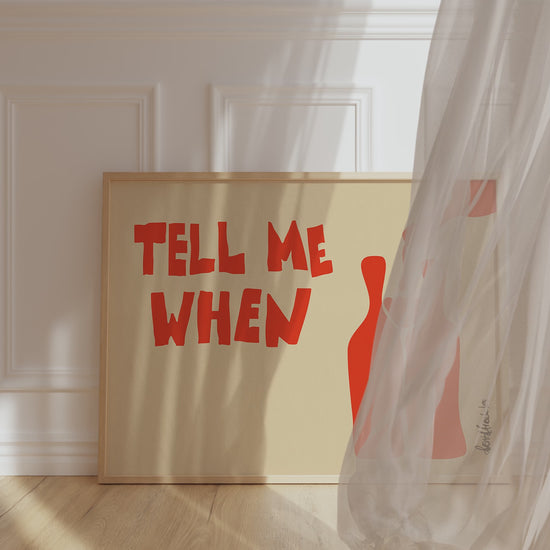 A framed picture leaning against a kitchen wall, beige background and bold orange letters reading "Tell Me When", while wind is blowing window curtain inside.