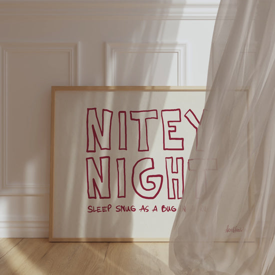 A framed picture leaning against a bedroom wall, beige background and bold red letters reading "Nitey Night", while wind is blowing window curtain inside.