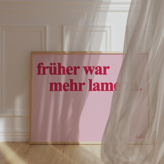 Ein gerahmtes Bild, das gegen ein Wand lehnt, zeigt den deutschen Text "Früher war mehr Lametta", ein Zitat von Loriot, in roter Schrift auf pinkem Hintergrund. Ein Vorhang weht vor den Rahmen durch Wind.