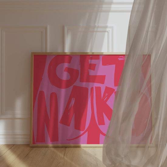A framed picture leaning against a bathroom wall, red background and bold pink letters reading "Get Naked" with a butt cheek drawing in its center.