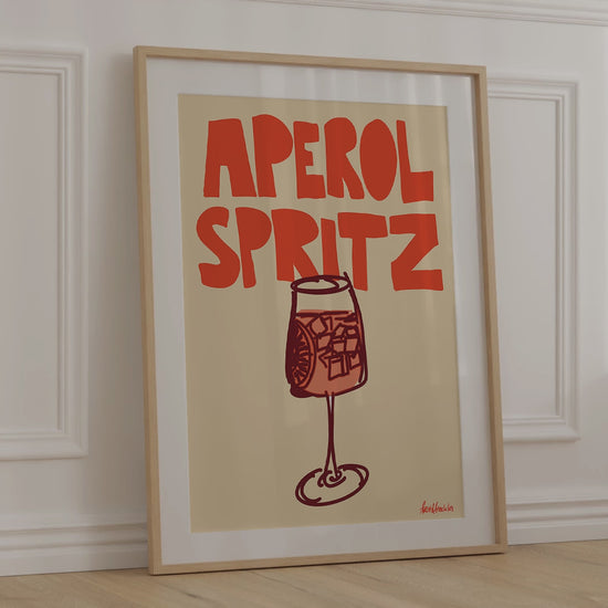 A framed picture leaning against a bedroom wall, beige background and bold orange letters reading "Aperol Spritz"