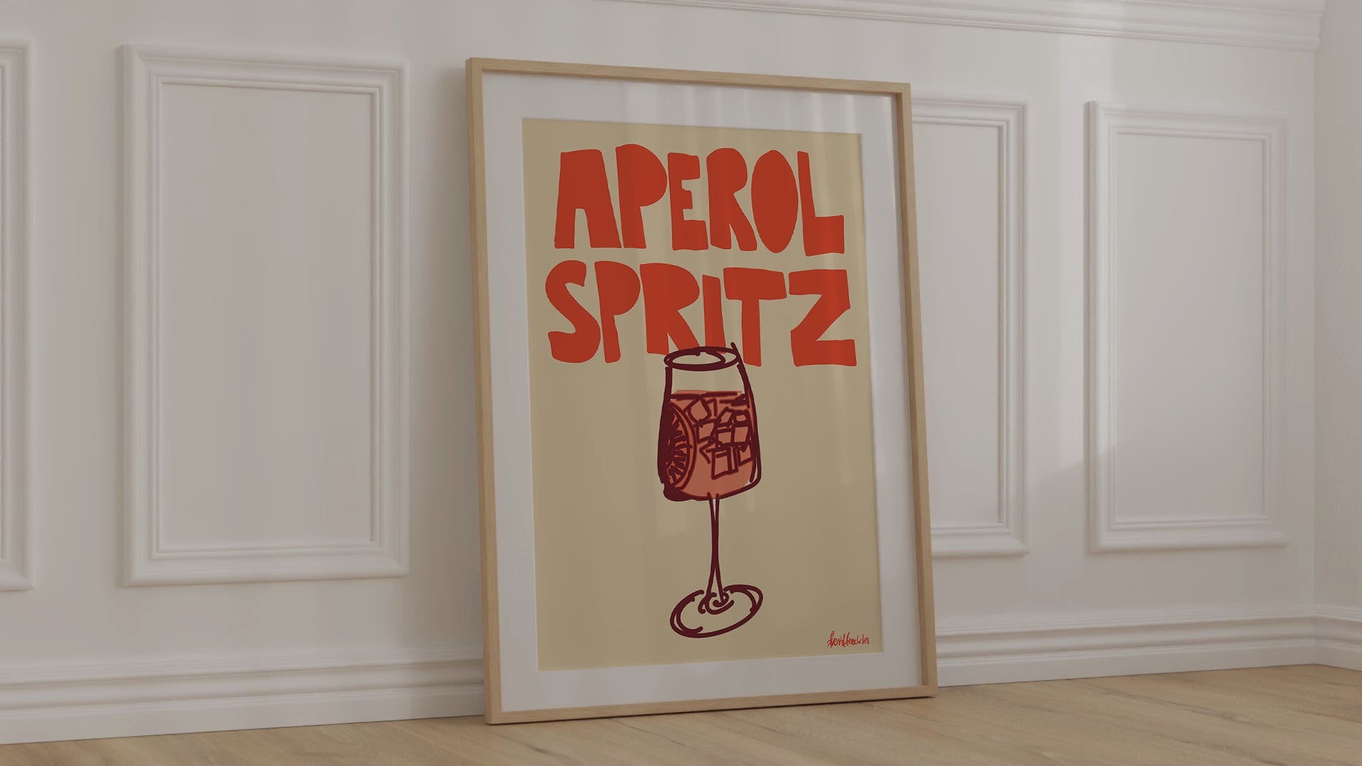 A framed picture leaning against a bedroom wall, beige background and bold orange letters reading "Aperol Spritz"