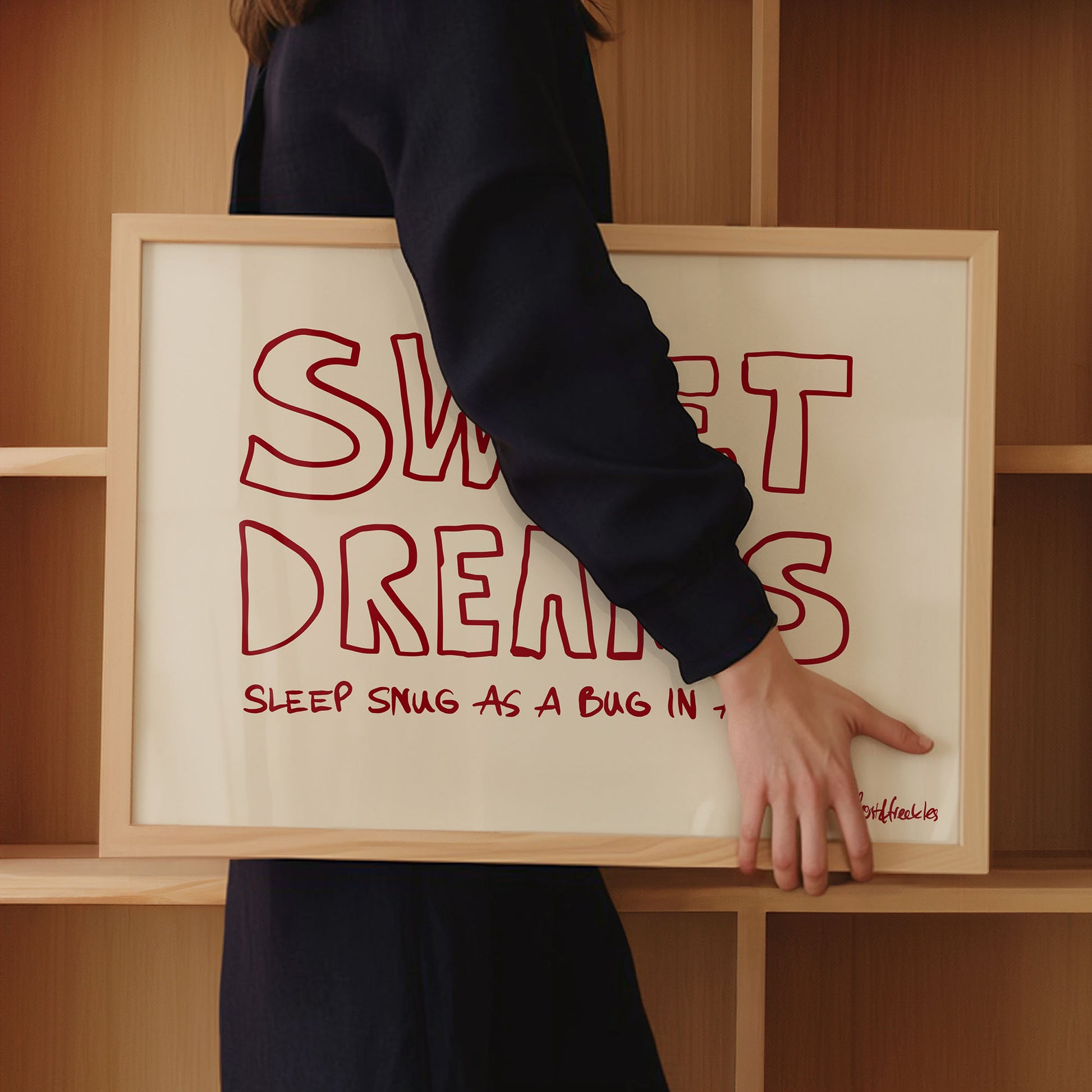 A woman in dark clothing holds a framed pink print with red text"Sweet Dreams" in front of wooden bedroom shelves.
