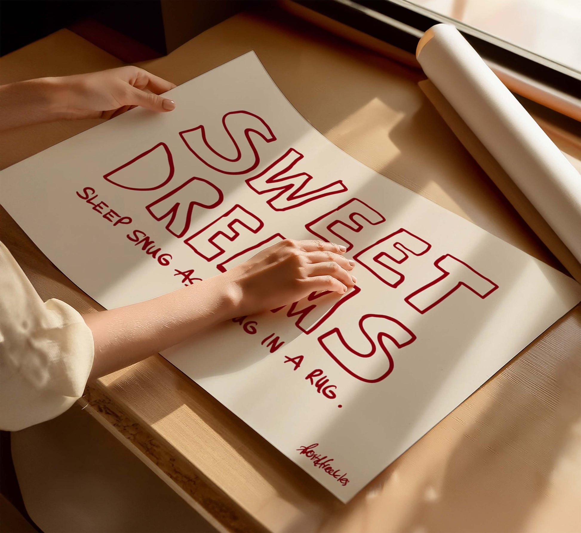 A woman holding a beige poster with the phrase “Sweet Dreams", in bold red text, on a wooden table near a window with sunlight. 
