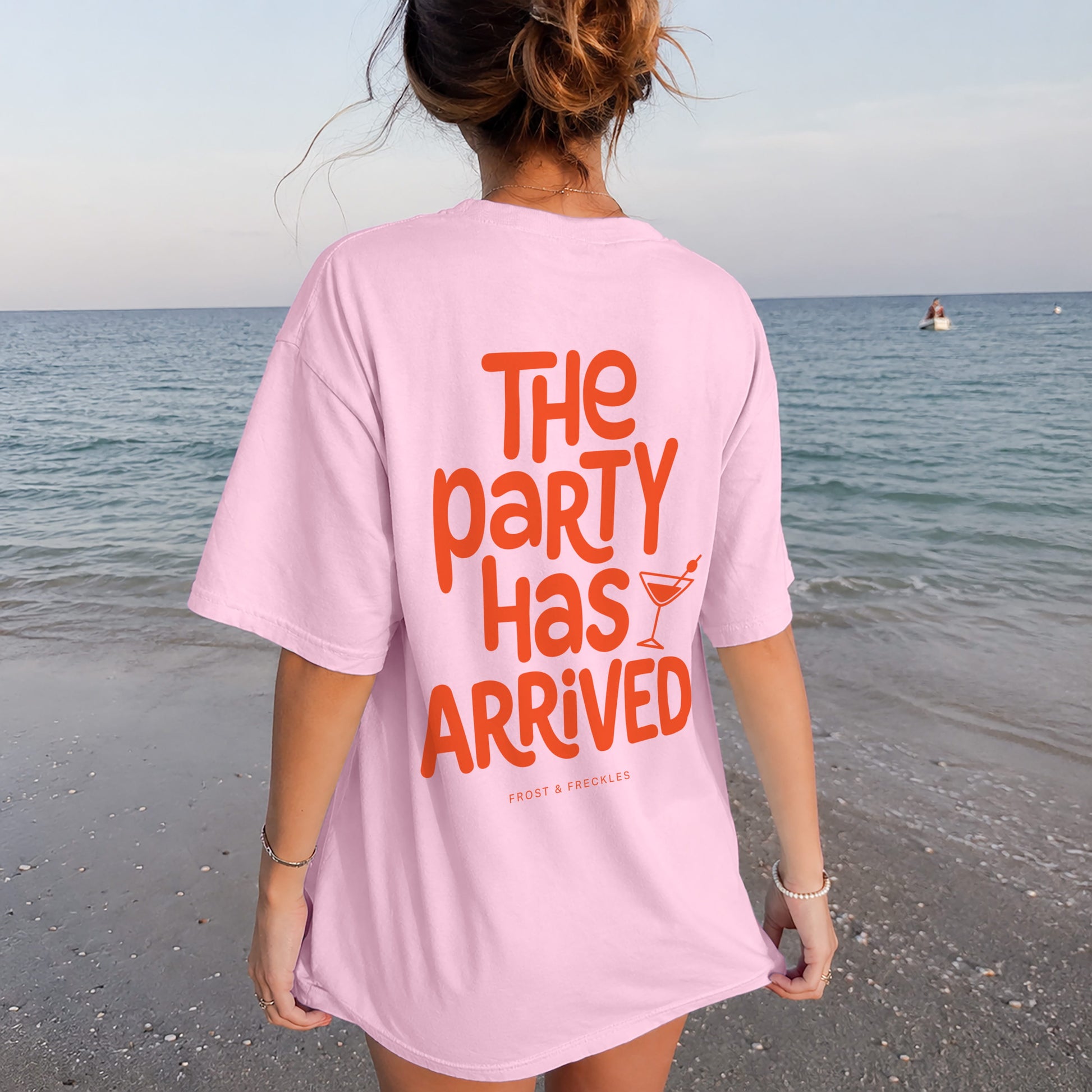 Frau von hinten vor einem Oldtimer-Van am Meer mit dem rosa Oversized T-Shirt und orangefarbenem Aufdruck "THE PARTY HAS ARRIVED", im exzentrischen Kaulitz & Kaulitz Party-Stil.