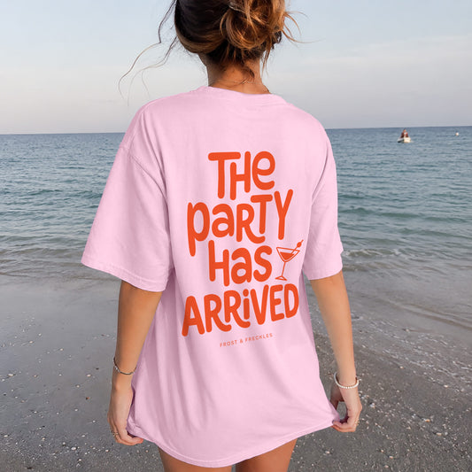 Frau von hinten vor einem Oldtimer-Van am Meer mit dem rosa Oversized T-Shirt und orangefarbenem Aufdruck "THE PARTY HAS ARRIVED", im exzentrischen Kaulitz & Kaulitz Party-Stil.