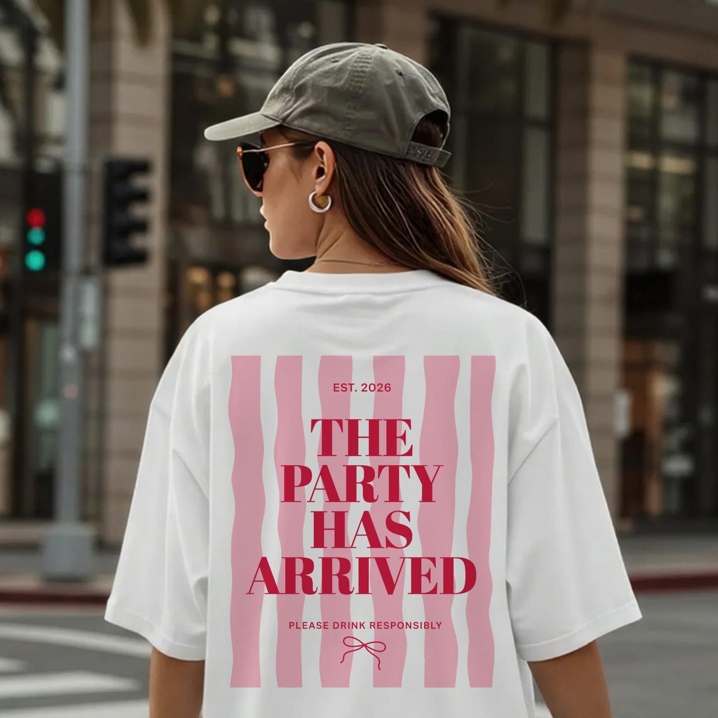 Frau von hinten vor einer Downtown Stadtkulisse mit dem weißen Oversized T-Shirt und pinken Aufdruck "THE PARTY HAS ARRIVED", im exzentrischen Kaulitz & Kaulitz Party-Stil.