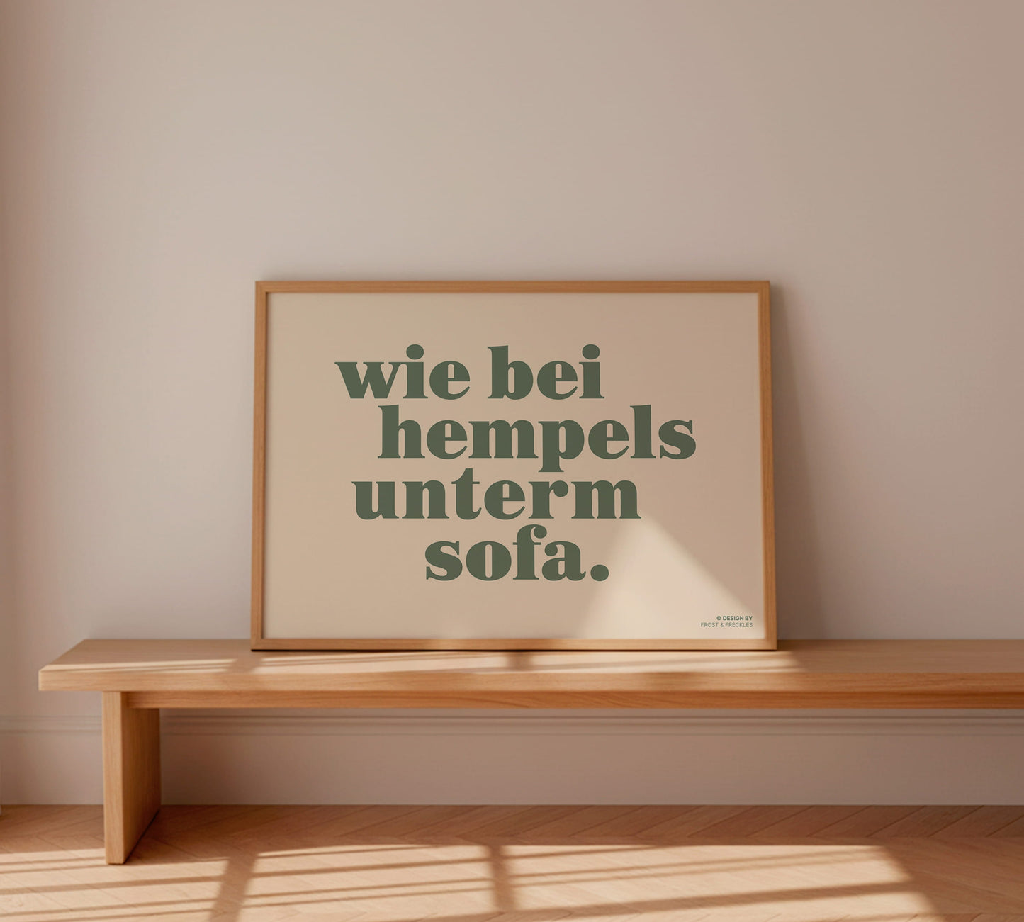 Gerahmtes Wandbild "Wie bei Hempels unterm Sofa" in einem hellen Holzrahmen auf einer modernen Holzbank lehnend.